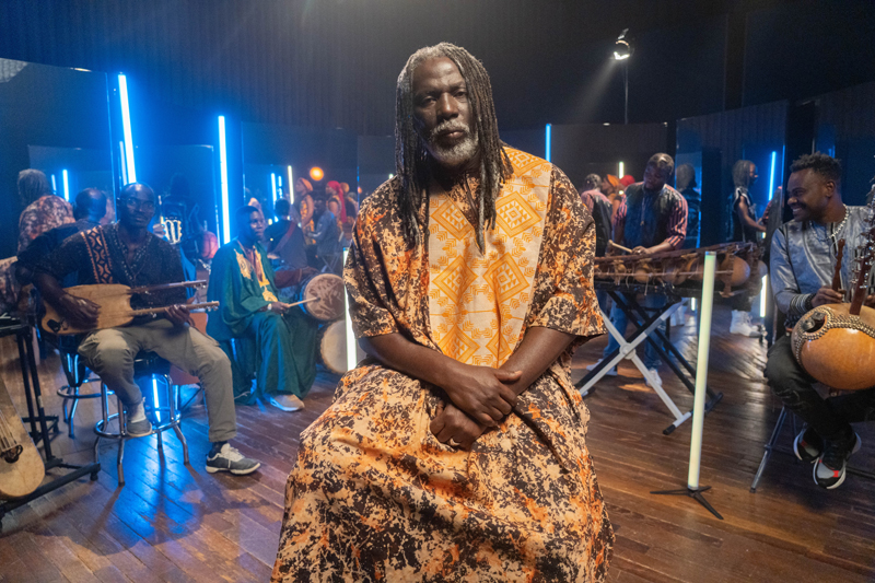 Tiken Jah Fakoly - Photo by Youri Lenquette. This photo features Tiken Jah Fakoly seated in the center of a musical setup. He exudes a commanding yet serene presence, dressed in a flowing, traditional African garment adorned with warm orange and earth-toned patterns. His long, gray-tinted dreadlocks frame his face. Behind him, an ensemble of musicians surrounds the space, playing traditional West African instruments such as koras, balafons, and ngonis.