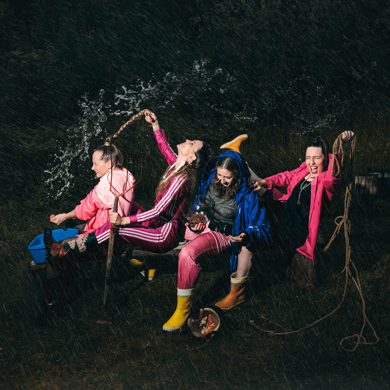 Tuuletar - Heponi mun (Mój koń nieborak) single artwork. A rain-soaked, high-energy scene shows Tuuletar's four women in bright outdoor gear laughing and flinging water as they sit on a bench in the dark. Each holds a rope or playful prop.
