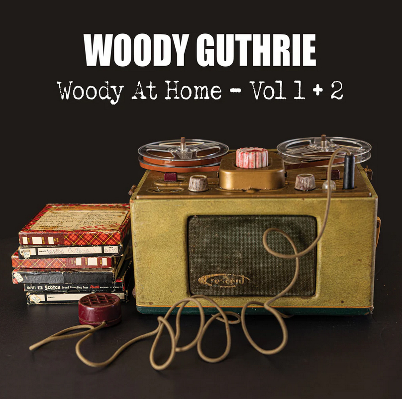 The album cover for Woody At Home – Vol 1 + 2 by Woody Guthrie features a vintage reel-to-reel tape recorder with tangled wires in front, placed beside a small stack of boxed tapes. The background is dark, making the bold white text of Guthrie’s name and album title stand out clearly above the retro equipment.