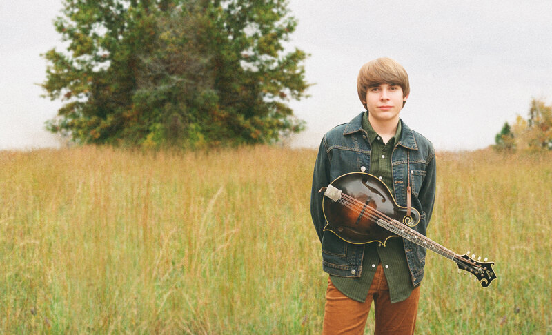 Wyatt Ellis standing in a field with his mandolin strapped.