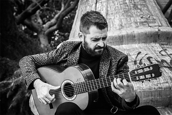 This is a black-and-white photo of flamenco guitarist José Almarcha. He is sitting outdoors, intensely focused while playing a flamenco guitar. He wears a patterned jacket over a dark turtleneck, with a natural backdrop enhancing the image's depth.