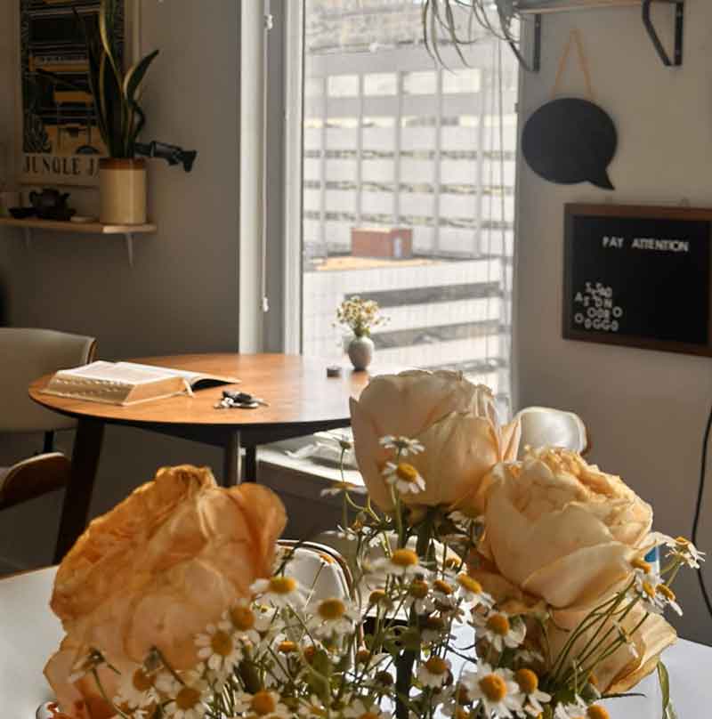 The cover of Mckey — Most of the Blame shows a warmly lit apartment interior with a vase of pale roses and daisies in the foreground. Behind them, a round wooden table holds an open book and keys near a window overlooking a city building. On the wall, a black letter board spells out “PAY ATTENTION.”