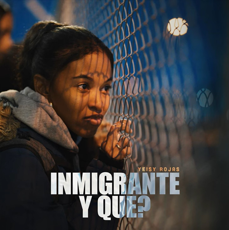 The single cover for Inmigrante Y Qué? by Yeisy Rojas features a young woman gazing pensively through a chain-link fence at night. Dramatic lighting casts the fence’s shadow across her face.
