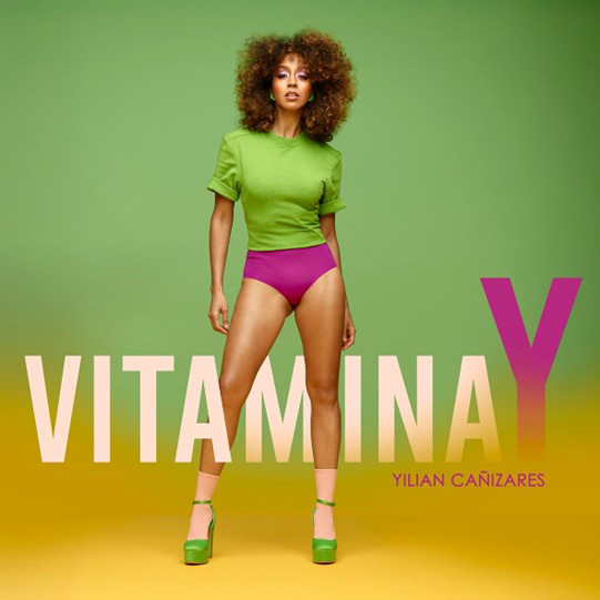 Yilian stands confidently against a green-to-yellow backdrop, styled in a bright green top, magenta bikini bottom, and matching platform heels. Pastel lettering spells out Vitamina Y, with the large final “Y” shifting from pink to purple.